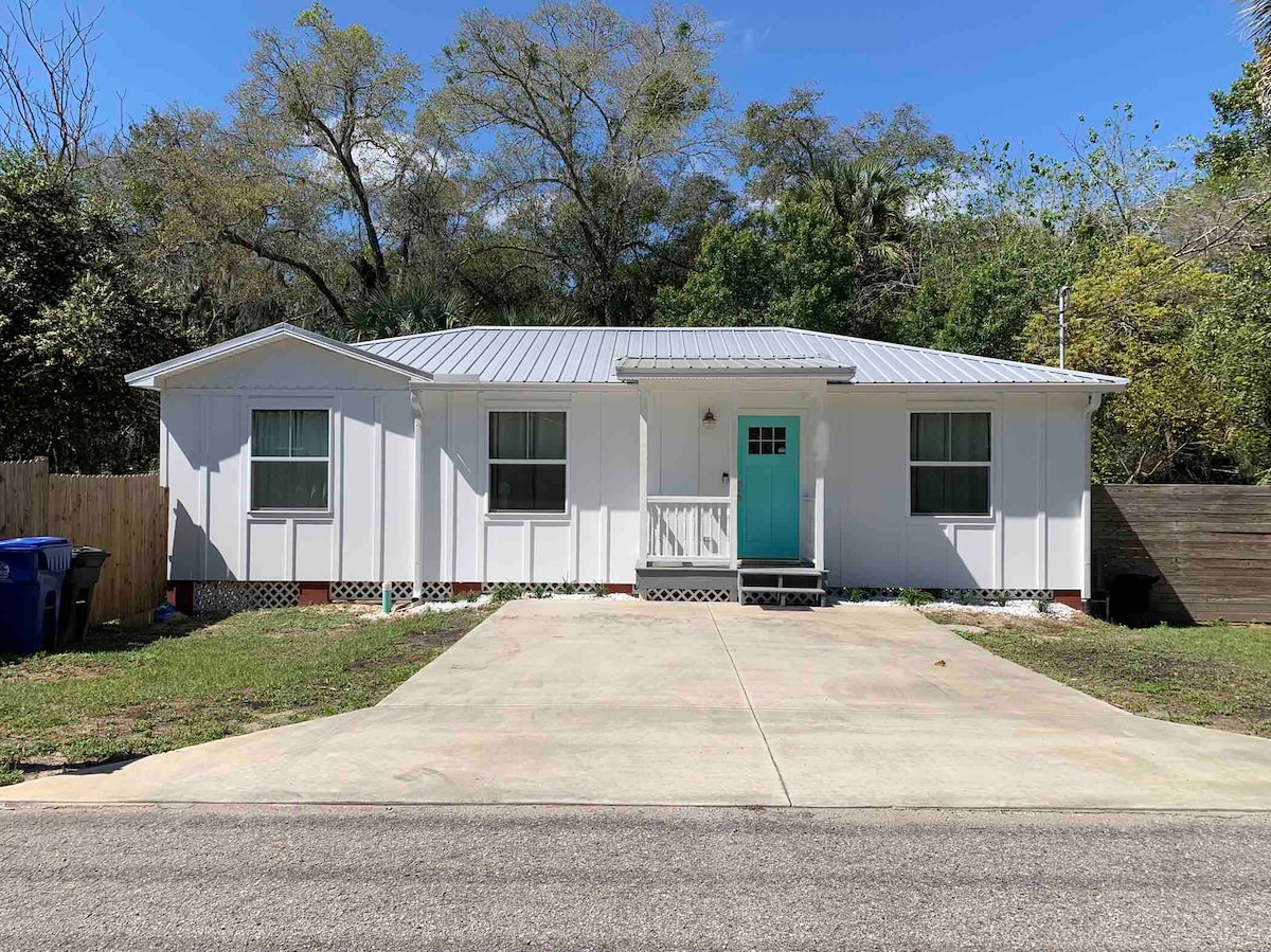 A single-story cottage is presented, featuring a light-colored facade with a contrasting teal front door. The driveway accommodates two vehicles, while windows on either side allow natural light to enter. A small patio area is visible at the entry, surrounded by a verdant lawn.