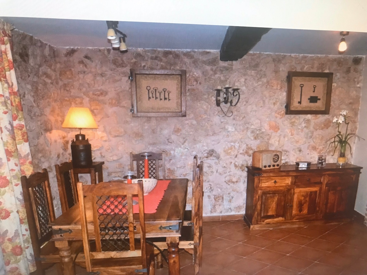 A rustic dining area is observed, featuring a wooden table surrounded by matching chairs. Stone walls are visible, adorned with framed key art. A soft-lit lamp sits on a side table, while a wooden sideboard displays decorative items and a potted plant.