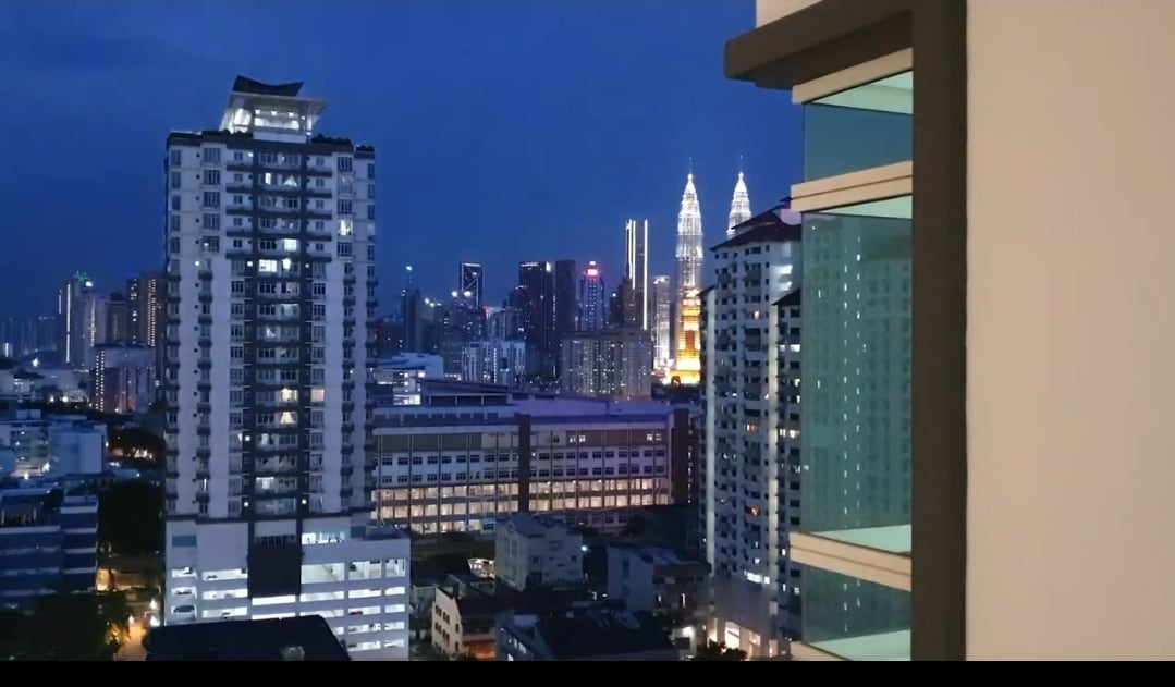 The image captures a cityscape at night showcasing a view of illuminated skyscrapers, including the distinctive twin towers. The city lights create a vibrant atmosphere, while a modern building's balcony provides a glimpse of the bustling urban setting framed against a twilight sky.