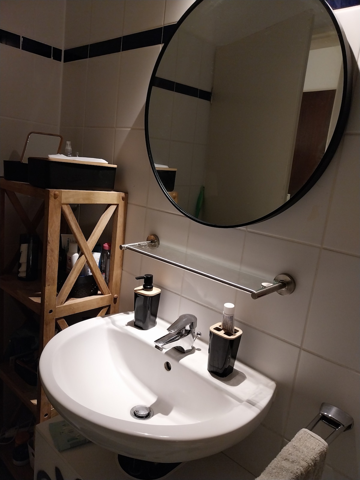 A modern bathroom is presented with a round mirror above a white sink. Dark bottles and a towel are neatly arranged on a chrome rail. A wooden shelf displays various toiletries, adding an organized touch to the space.