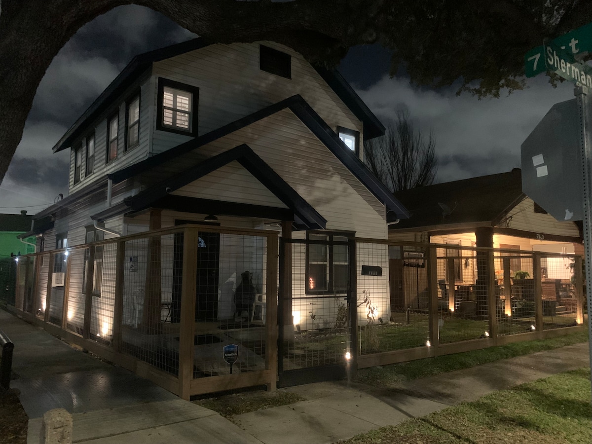 A two-story house is presented at night, with outdoor lighting highlighting its architectural features. A fenced yard surrounds the property, and a covered porch is visible on the right, providing shaded seating. The street sign is partially shown, indicating the location.