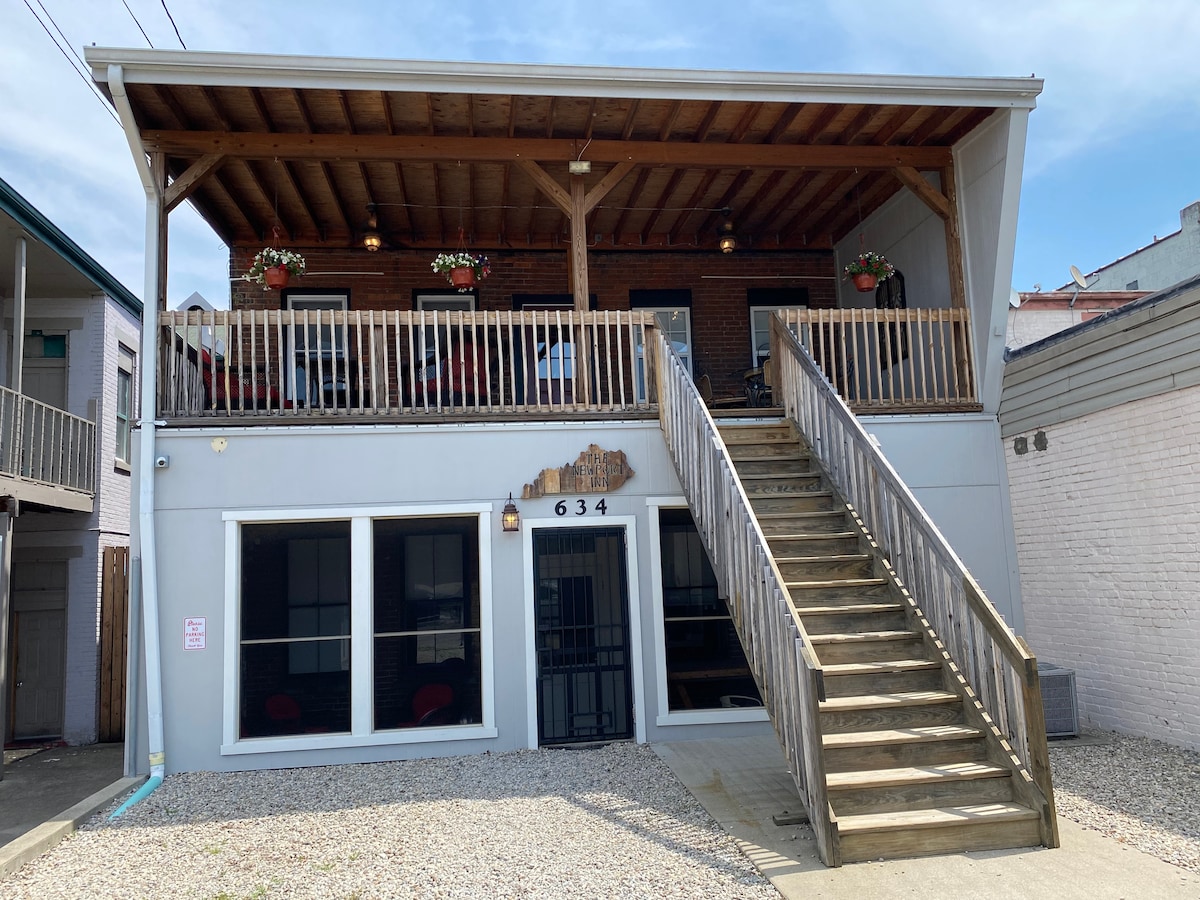 A two-story building features a welcoming entrance with a set of stairs leading to a spacious deck adorned with potted plants. Large windows on the ground floor provide natural light, while the upper deck is furnished for relaxation.