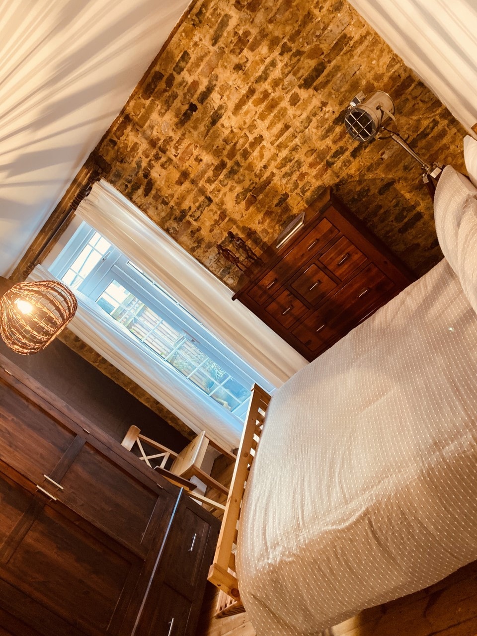 A cozy bedroom features exposed brick walls and natural light filtering through a large window. A spacious bed is accompanied by a wooden dresser and a bedside lamp. Light-colored curtains add a soft contrast to the room's warm tones.
