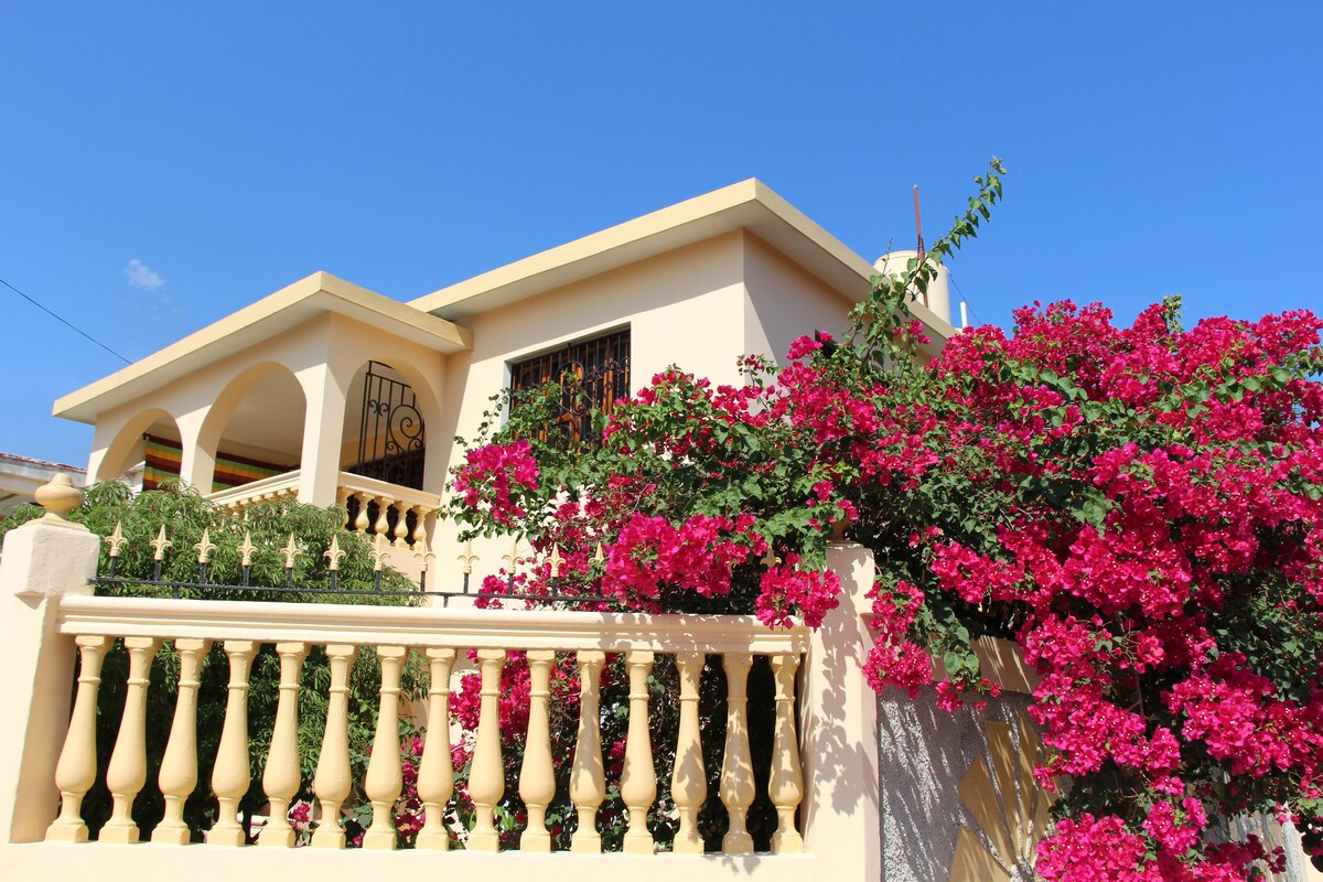 A colonial-style house features a welcoming façade, adorned with vibrant pink bougainvillea cascading over a decorative balcony. The structure is framed by a clear blue sky, enhancing the charm of the entrance and surrounding greenery.