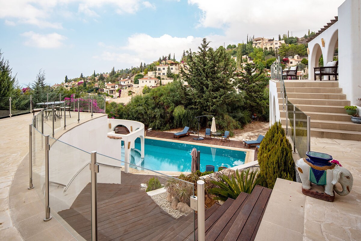 The outdoor area features a spacious pool surrounded by lounge chairs, with a view of the hillside and residential homes. A gentle slope is visible, leading from the pool to a patio area. Lush greenery frames the scene, enhancing the serene ambiance.