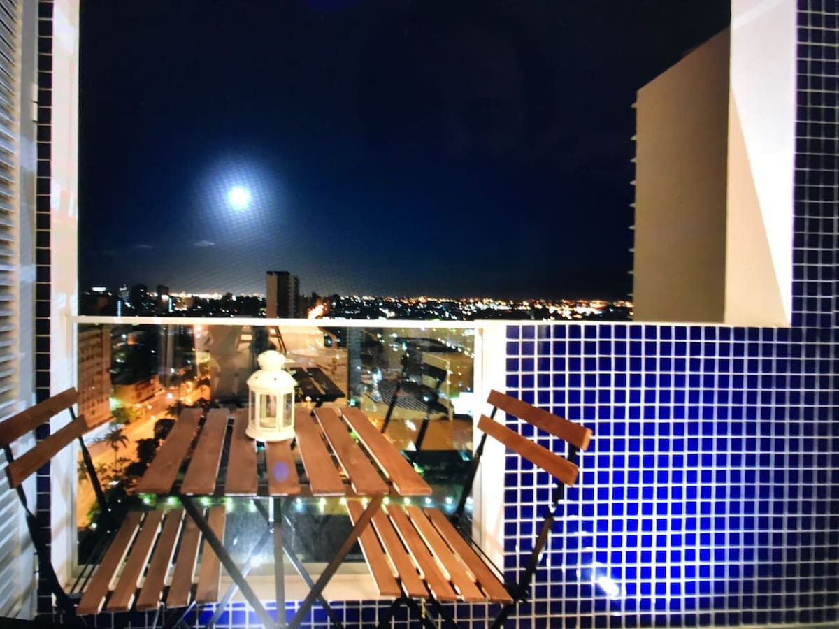 An outdoor balcony is depicted, featuring a compact wooden table with two chairs. A lantern rests atop the table, with a panoramic view of a city skyline illuminated at night. The walls are adorned with blue tiles, contributing to the overall aesthetic.