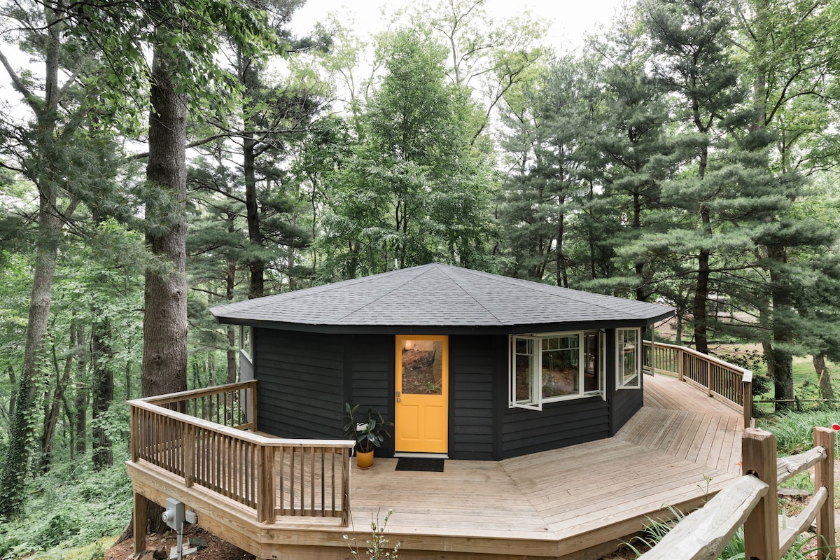 The unique 13-sided round home is surrounded by lush greenery, with a spacious wooden deck that extends around the exterior. Large windows allow for abundant natural light, while a vibrant yellow front door adds a pop of color against the dark wood siding.
