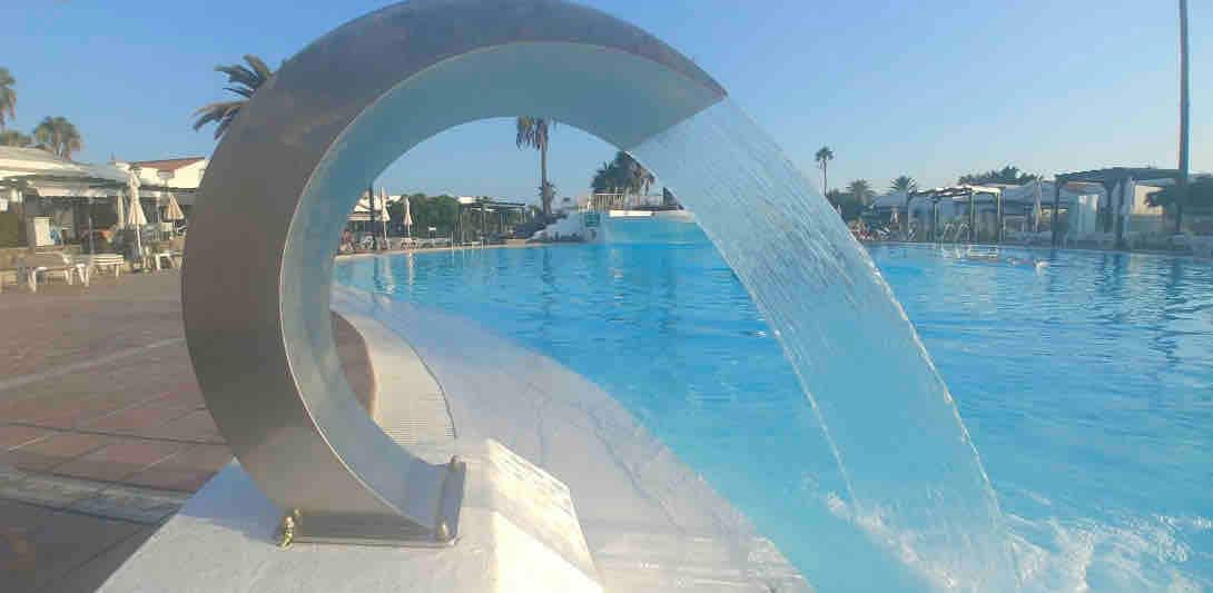 A sleek, contemporary water feature sends a gentle cascade into a large swimming pool, reflecting the bright blue sky. Surrounding palm trees and lounge chairs indicate a resort-style ambiance, contributing to a relaxed atmosphere for guests to enjoy leisure activities.
