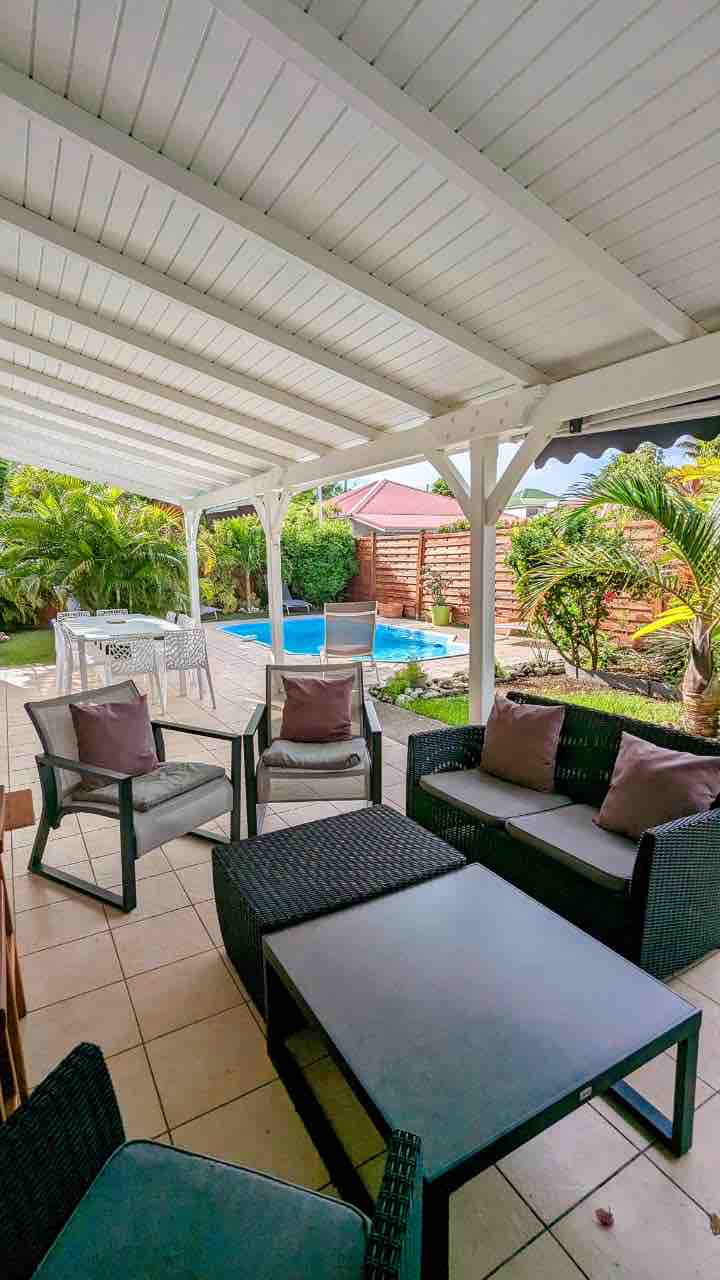 An outdoor seating area is situated under a protective roof, featuring dark wicker chairs and a coffee table. An inviting pool can be seen in the background, surrounded by lush greenery and a sunlit patio area with additional seating and vibrant landscaping.