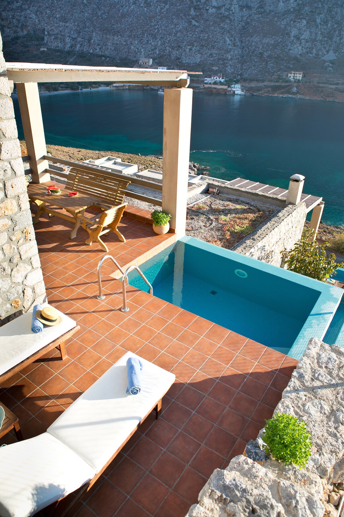 A private terrace showcases a small swimming pool next to a tiled area with two loungers and a matching table with chairs. The setting offers views of the sea and rocky landscape, complemented by potted greenery along the edges.