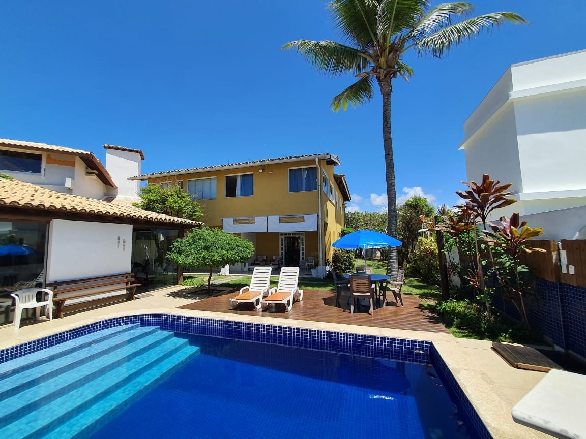 A colorful two-story house is shown in a sunny garden setting, featuring a swimming pool with a gradient blue surface. Lounge chairs are positioned poolside under the shade of a large palm tree, while a vibrant blue umbrella provides additional shade. Lush green trees surround the property.
