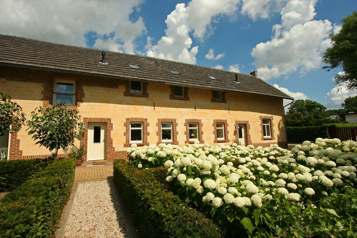 A spacious farmhouse exterior is surrounded by lush greenery and blossoming flowers. The building features a warm brick facade, with multiple windows inviting natural light. A pathway lined with bushes leads to the entrance, framed by the vibrant garden.