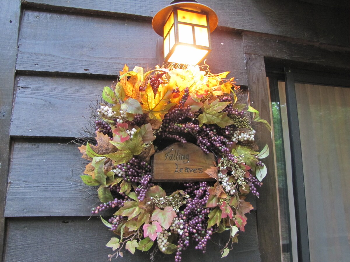 A decorative wreath made of autumn foliage and berries adorns the entrance, featuring a small wooden sign that reads 'Falling Leaves.' A lantern above the wreath softly illuminates the area, highlighting the vibrant colors of the leaves.