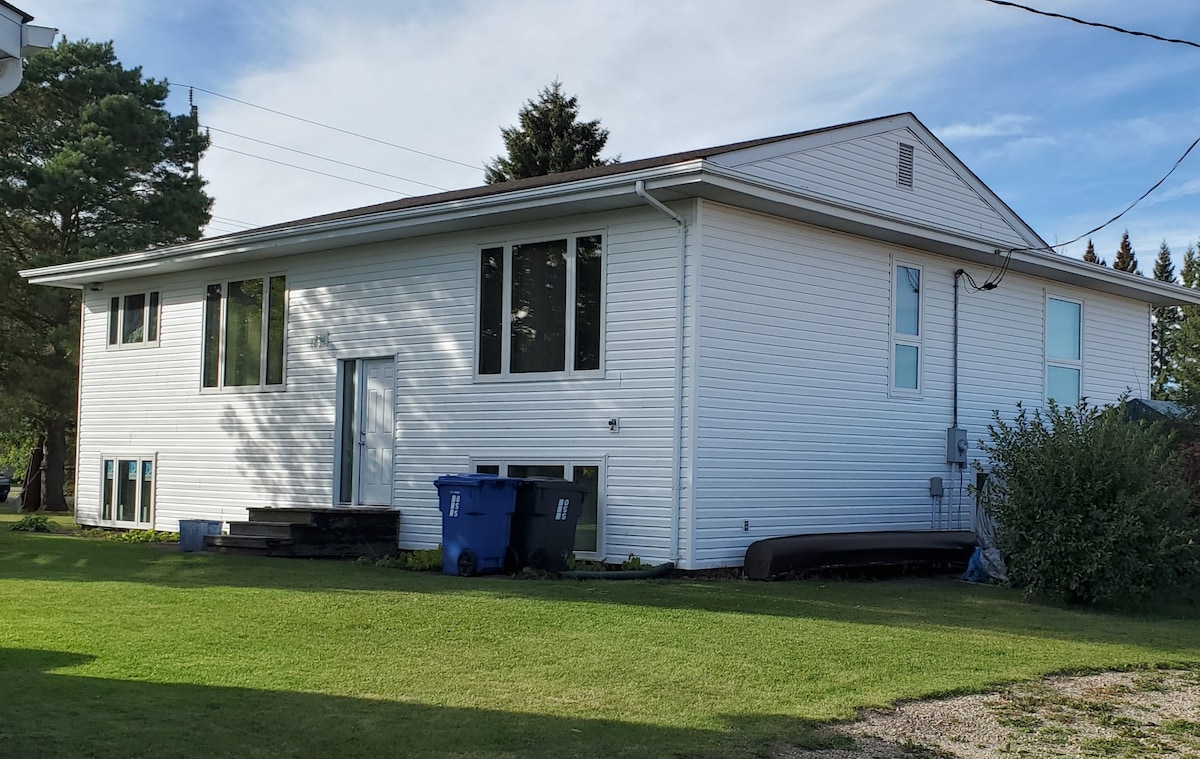 The exterior of a modern home is displayed, featuring a light-colored siding and a well-maintained lawn. Large windows allow natural light to enter, while a set of stairs leads to the main entrance. A blue recycling bin and a water tank are visible nearby.