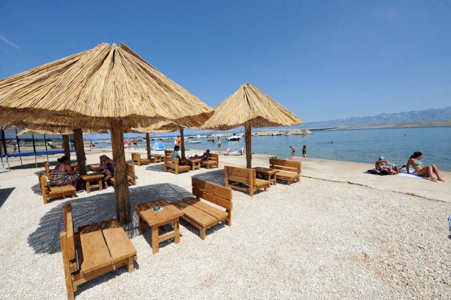 Beach huts with thatched roofs provide shaded seating near the water's edge. Wooden tables and benches are arranged on a pebble-covered surface. Calm blue waters and distant mountains create a serene backdrop, with visitors enjoying the sun and water activities.
