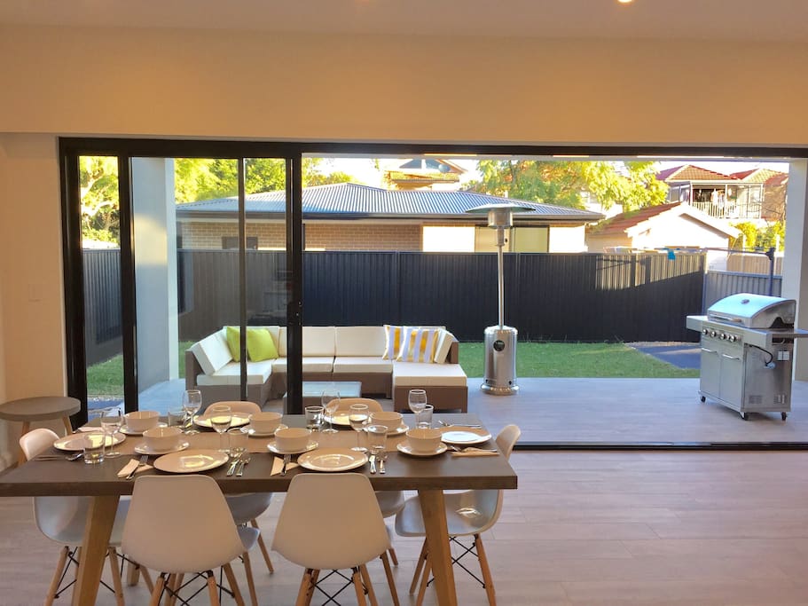 The spacious open-plan living and dining area leads straight through a set of floor to ceiling glass doors onto a tiled entertainer's terrace equipped with outdoor lounge, gas heater, and four-burner barbecue