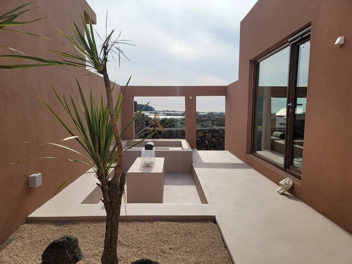 A serene outdoor space features smooth, light-colored concrete surfaces and a central area for relaxation. A small tree adds a touch of greenery, while a view of the distant sea and nearby land can be seen through large glass doors, creating an inviting atmosphere.