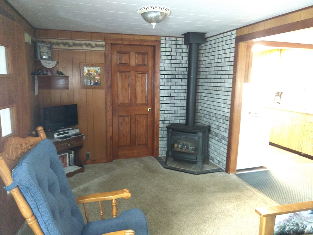 A cozy living area features wood-paneled walls and a warm carpet. A rustic rocking chair is positioned near a freestanding wood stove, while a small television sits on a shelf. Light from the adjoining kitchen illuminates the space, creating a comfortable atmosphere.