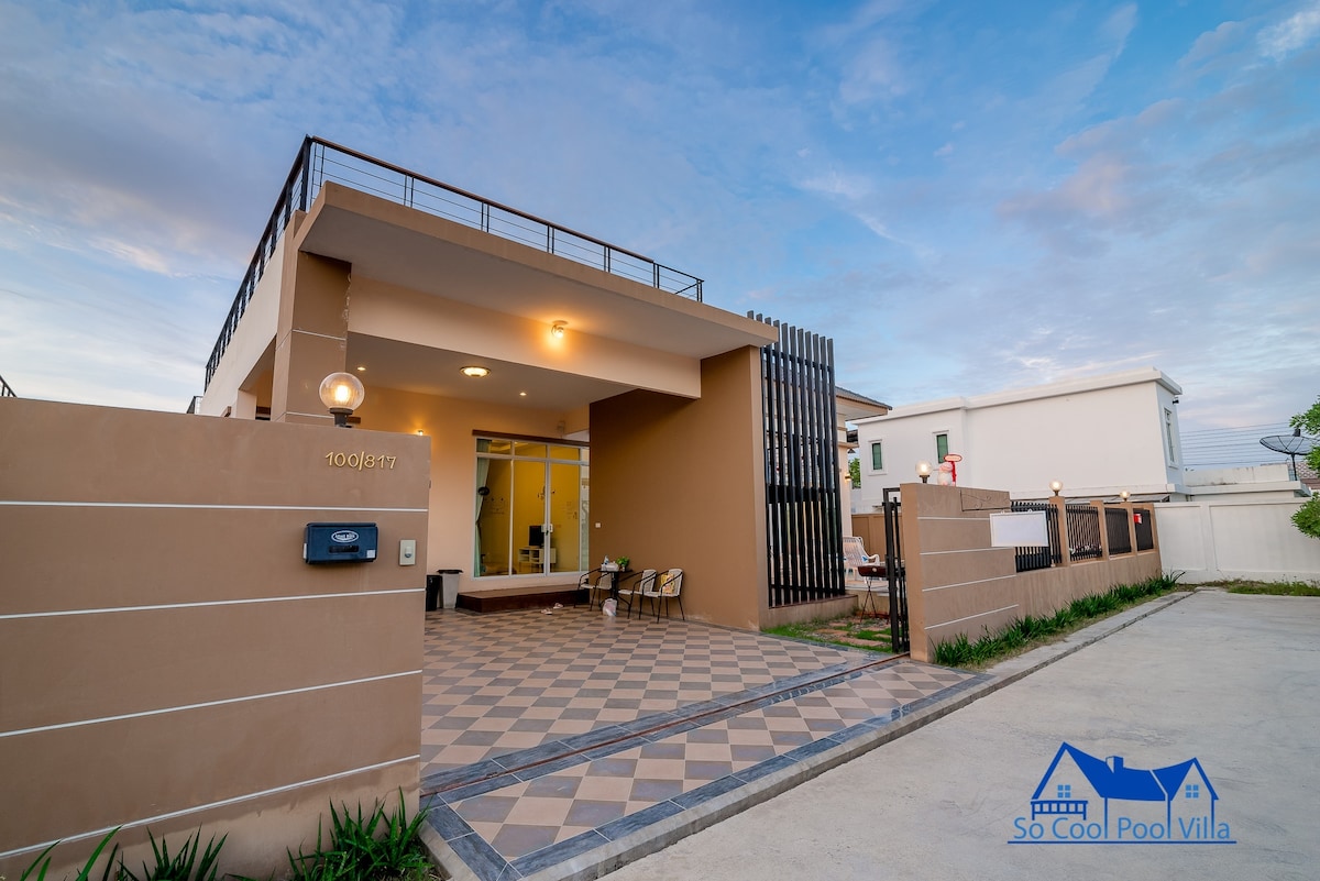 The exterior of the house features a contemporary design with a spacious entrance. A checkered pattern is visible on the driveway, leading to a well-lit patio space. Surrounding greenery and modern lighting add to the inviting facade.