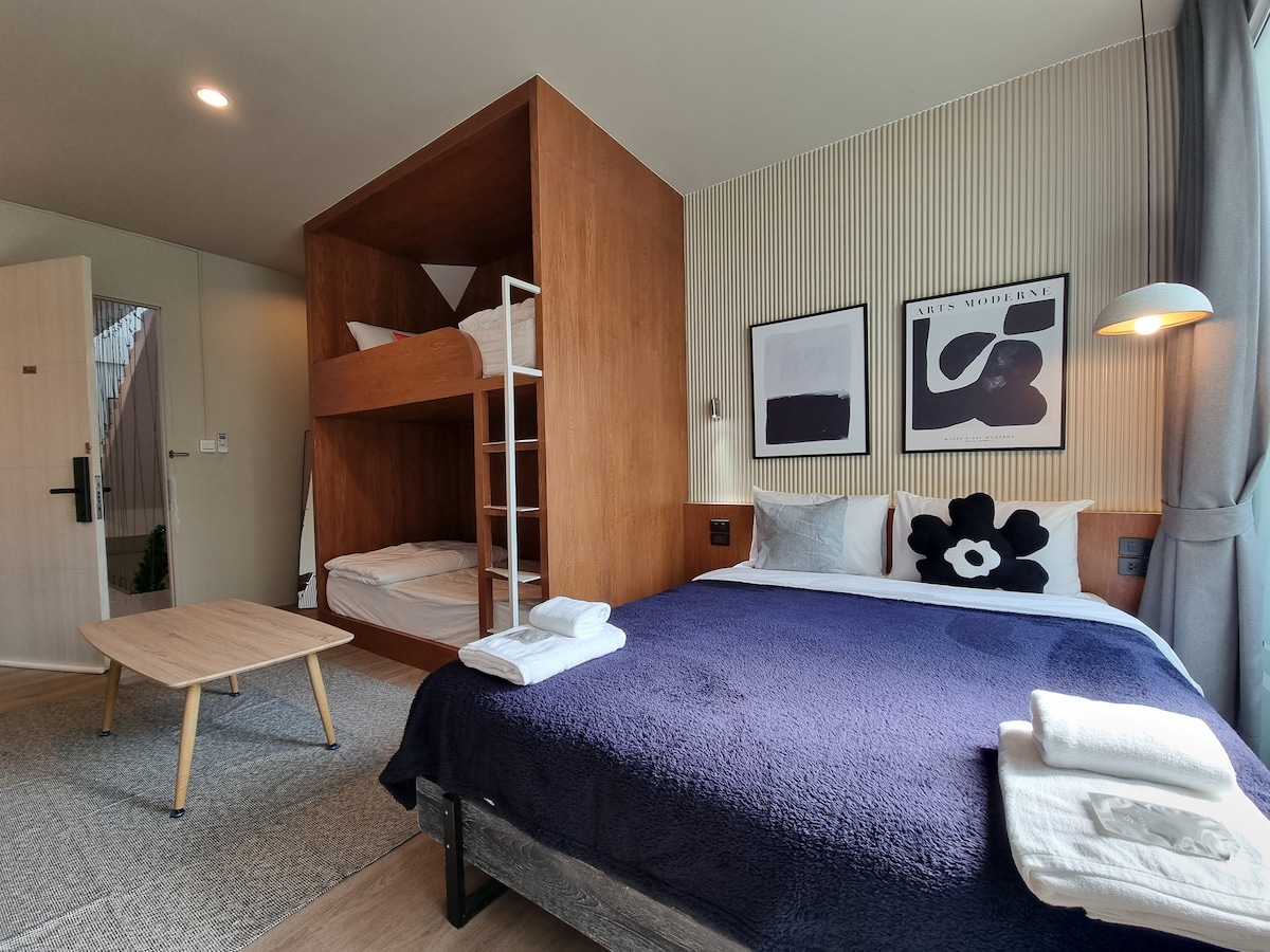 A comfortable sleeping area is showcased with a spacious bed covered in a navy blanket, and a cozy bunk bed positioned against a wooden accent wall. A small wooden table stands nearby, complemented by framed art on the wall and soft natural light filtering through the window.