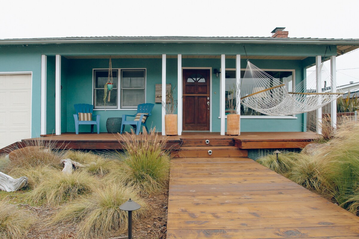 The front porch features a wooden walkway leading to a welcoming entrance. Two blue chairs are positioned on the porch beside a woven hammock. The entrance is framed by white columns, and a decorative stone element is also visible amidst the grassy landscaping.
