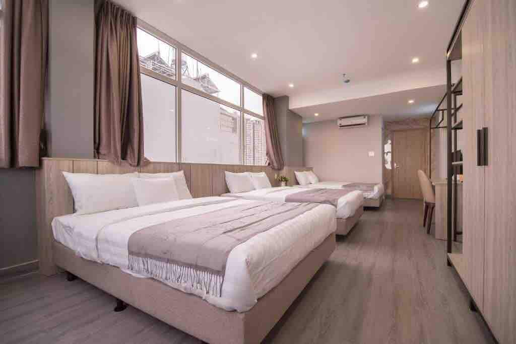 A modern room features two spacious beds with neutral bedding and a light grey throw blanket. Large windows allow natural light to fill the space, while elegant curtains complement the contemporary design. A small dining area is visible in the background.