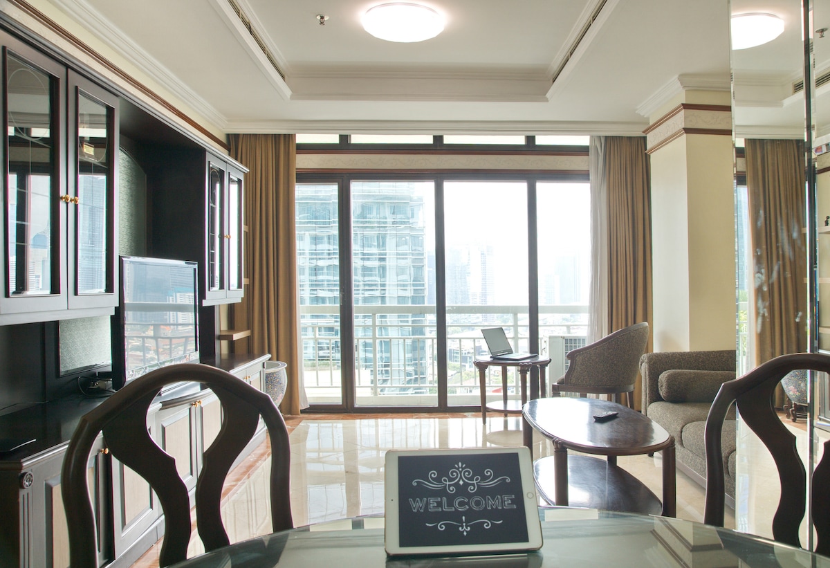 A bright living area is presented with large glass windows, offering views of the city skyline. Elegant furnishings include a round dining table and a comfortable sofa, enhancing the sophisticated ambiance with marble flooring reflecting natural light.