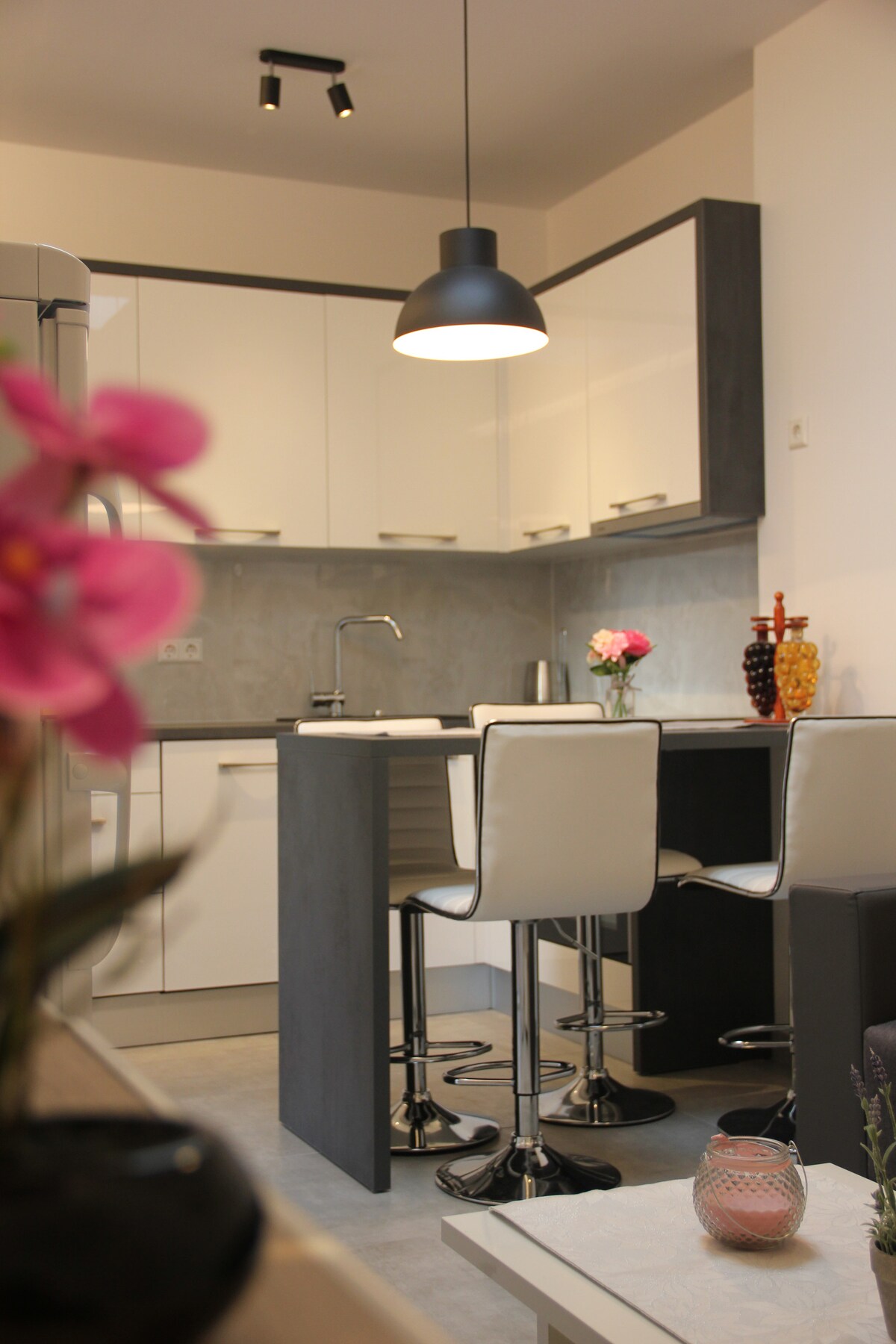 A modern kitchen is featured, equipped with sleek white cabinetry and a gray accent wall. Two high stools sit at a compact bar area, illuminated by a pendant light. Decorative items, including a vase with flowers and colorful glass objects, enhance the inviting atmosphere.