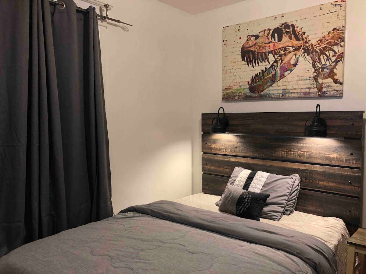 A cozy sleeping area features a wooden bed frame with a modern T-Rex artwork above the headboard. Soft gray bedding and a decorative black cushion enhance the space, while dark curtains provide privacy and control over natural light.