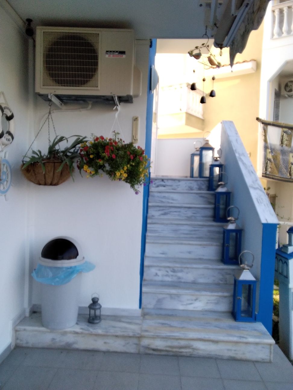 A bright and inviting stairway is seen, featuring marble steps and decorative blue railings. Potted flowers hang near the top, while an air conditioning unit is mounted on the wall. Lanterns line the staircase and a waste bin is positioned nearby.