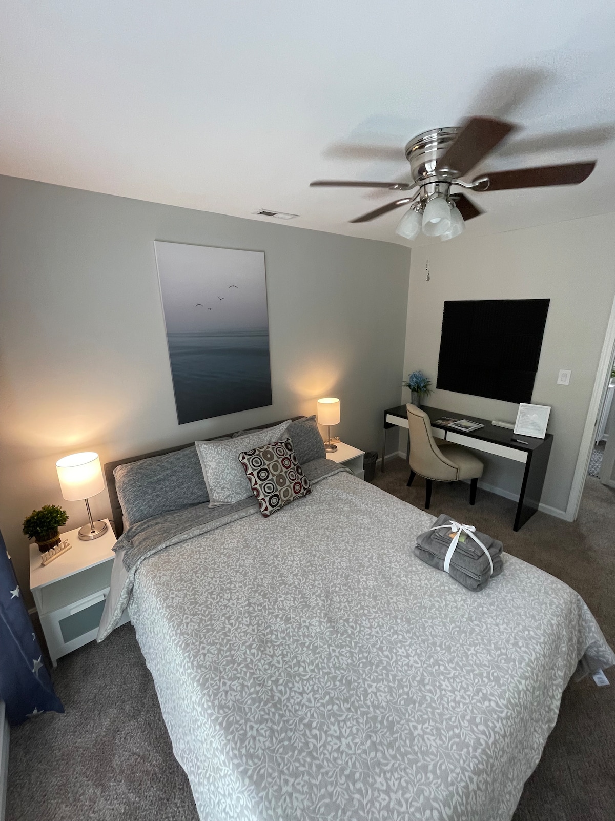 A calming bedroom is presented with a spacious bed adorned with a decorative gray and white blanket. A wall-mounted television is visible above a desk and chair. Two bedside tables are flanked by lamps, while a large artwork depicting a serene landscape hangs on the wall.