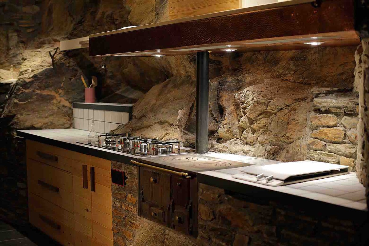 A kitchen area is designed with a blend of natural stone and wooden cabinetry. A gas stove with multiple burners is situated along a countertop, complemented by under-cabinet lighting and a natural rock wall backdrop. A notepad and pen rest on the counter surface.