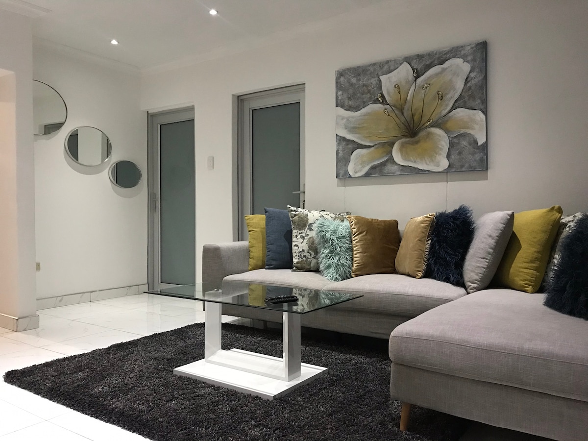 A light-filled living area features a sectional sofa adorned with colorful cushions in shades of yellow, teal, and blue. A glass coffee table sits on a textured dark rug, and a large floral artwork is displayed on the wall. Soft reflections are created by mirrored accents.