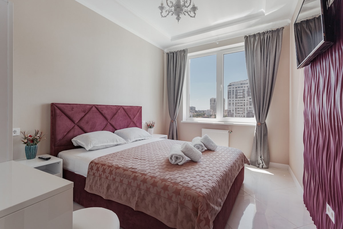 A well-lit bedroom is showcased, featuring a plush, upholstered bed with a decorative blanket. Soft pillows are neatly arranged on the bed. Floor-to-ceiling windows offer a view of the city skyline, complemented by elegant drapes framing the windows. A small decorative plant is visible on a nightstand.