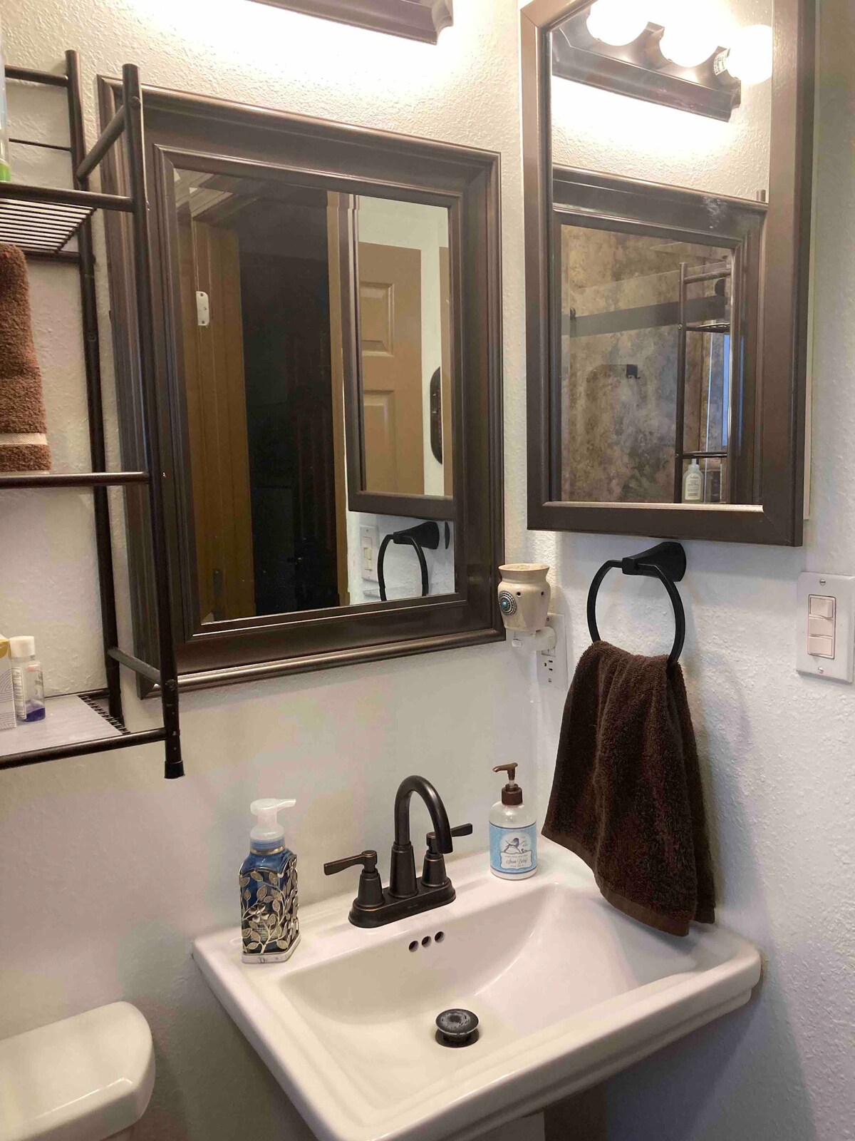 The bathroom features a compact sink with a modern faucet, complemented by two framed mirrors. Shelving is visible on the left, holding neatly arranged towels and toiletries. A towel hangs on a ring beside the sink, and a hand soap dispenser is positioned nearby.