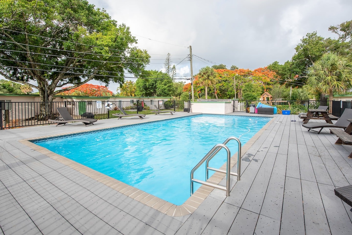 A large, outdoor saltwater pool is surrounded by a spacious deck with several lounge chairs. Lush trees and greenery provide natural shade, while vibrant flowers can be seen in the background. The pool area showcases inviting turquoise water and is protected by fencing.