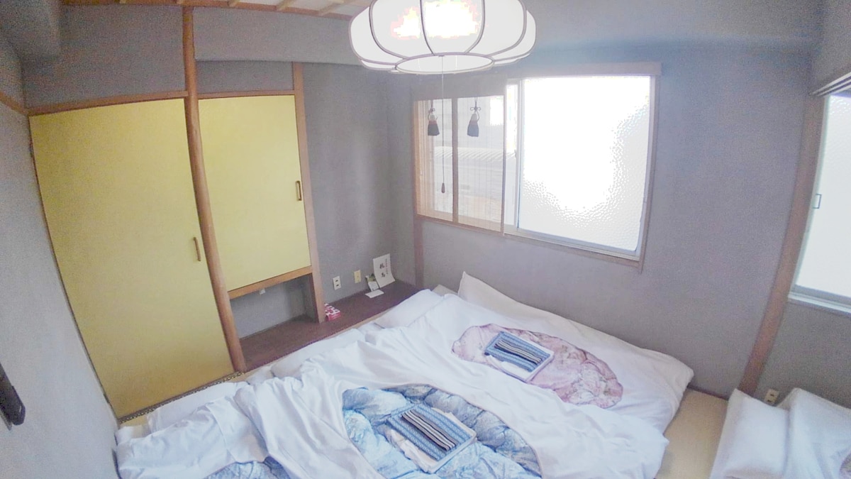 A spacious guest room is presented with traditional futons arranged on the floor, complemented by subtle pastel-colored patterns. Natural light filters through large windows, illuminating the pale walls and wooden accents. A minimalist workspace is visible, featuring a small desk and a yellow sliding door.