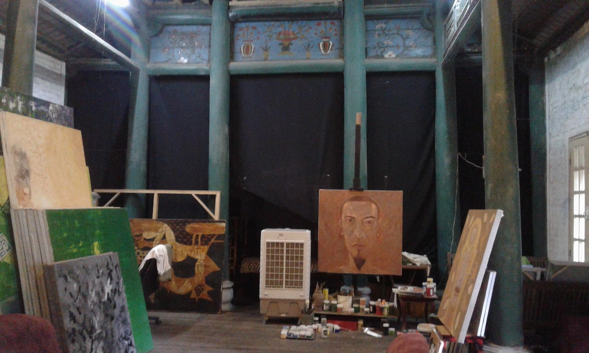 An artsy indoor space is characterized by tall green pillars and a ceiling adorned with decorative elements. Various canvases and art supplies are arranged around the room, with a portable air conditioner positioned against the wall. Dark curtains create a contrast against the bright color scheme.