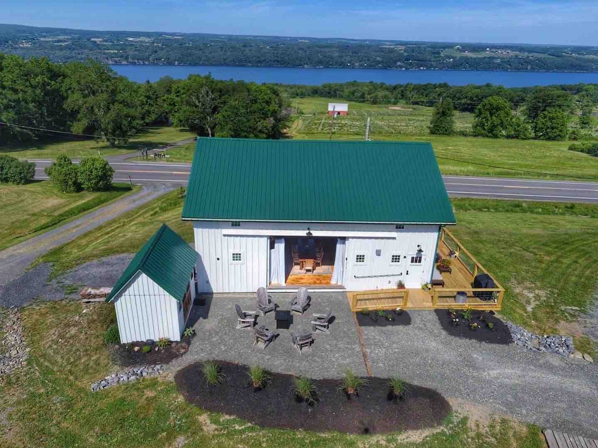 An aerial view captures the barn's unique green roof and white exterior. A spacious deck is visible, surrounded by a gravel area with seating arranged around a fire pit. Seneca Lake and lush countryside stretch into the distance, enhancing the tranquil setting.