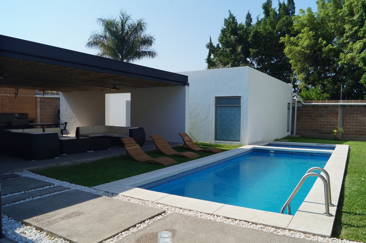 A modern pool area features a clear blue swimming pool surrounded by a well-maintained lawn. Two lounge chairs are positioned nearby, providing relaxation space. The shaded seating area offers a comfortable spot for outdoor gatherings, with a backdrop of lush greenery and a clean, minimalist facade.