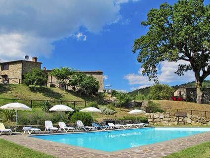 A refreshing swimming pool is surrounded by comfortable lounging chairs and shaded umbrellas. Lush green hills serve as a backdrop, featuring rustic stone buildings and trees. The scene captures a peaceful outdoor atmosphere under a partly cloudy sky.
