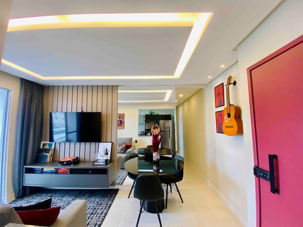 A modern living area is illustrated with a dining table surrounded by black chairs. A wall-mounted television is positioned above a sleek console. The kitchen area is visible in the background, complemented by artwork and a guitar displayed on the walls.