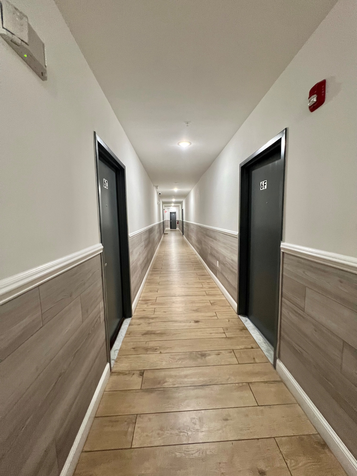 A well-lit hallway features a long wooden floor with light-colored planks. The walls are painted in a soft neutral tone, accented by a darker wood paneling halfway up. Dark doors are visible along either side, creating a symmetrical and inviting atmosphere.
