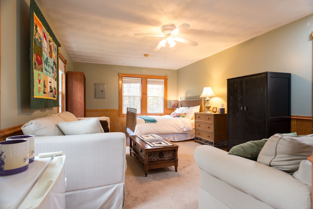 A spacious room features a comfortable bed, accompanied by a cozy seating area with two armchairs and a coffee table. A large dresser and a dark wardrobe provide ample storage. Soft natural light enters through the windows, illuminating warm tones throughout the space.