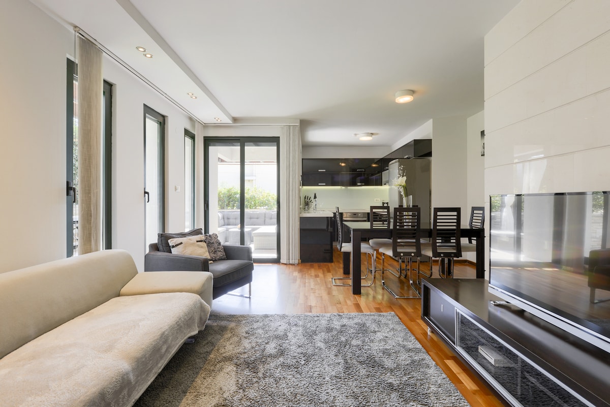A modern living area is showcased with large windows allowing natural light. A spacious sofa and an armchair are arranged over a soft rug. A dining table and kitchen are visible in the background, providing an open concept layout. A TV stands against a feature wall.