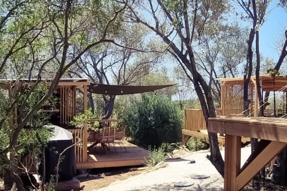 An outdoor space is framed by lush trees, showcasing a wooden deck with a dining area under a shaded canopy. A jacuzzi is positioned nearby, surrounded by natural elements. Pathways of gravel lead through the tranquil garden setting, enhancing the connection to nature.