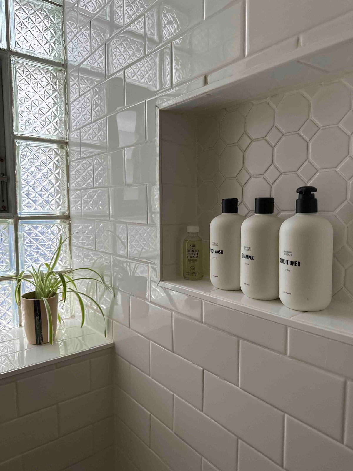 A well-lit bathroom features a niche with three neutral-toned bottles labeled as shampoo, conditioner, and body wash. A small potted plant adds a hint of green against the backdrop of white hexagonal tiles and glass block windows, enhancing the space's clean design.