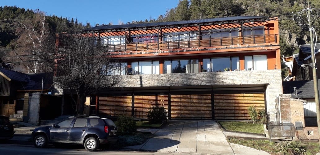 A modern building with a two-story structure features a combination of wood and stone elements. Large windows provide ample natural light, and a covered balcony offers outdoor space. The landscape includes trees and a well-maintained front yard adjacent to the parking area.