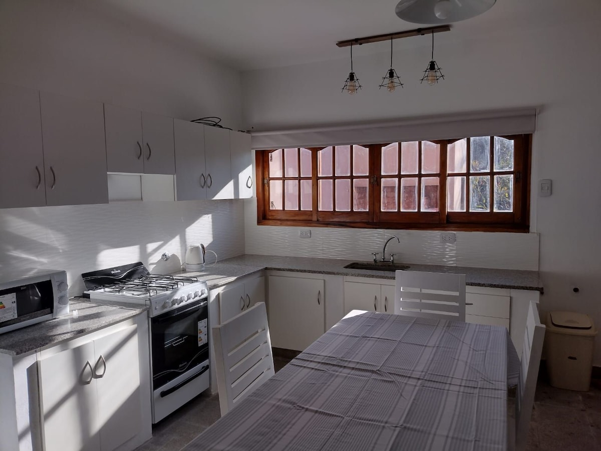 A bright kitchen is equipped with light-colored cabinets, modern appliances, and a spacious dining table. Natural light floods the area through large windows, illuminating the countertop and providing a welcoming cooking space.