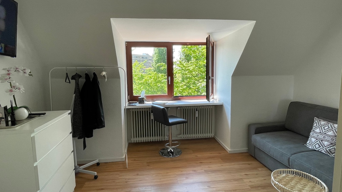 A bright room features a large window showcasing green views of surrounding trees. A sleek desk chair sits beside a simple desk, while a gray sofa offers comfortable seating. A clothing rack and a white dresser provide additional storage options.